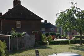 The image depicts the front yard of a residential house, viewed from the street, in Becontree, with a typical UK suburban setting. In the foreground, there are several cardboard boxes stacked and scattered on a paved driveway area, some partially open revealing items inside. Beside the boxes, a large wooden wardrobe with a dark finish and visible wood grain is being carried by two individuals, with one holding the top and the other supporting the base. The individuals appear to be engaged in a house removal activity, assisted by a trolley or dolly positioned nearby. To the right of the scene, a white removals van is parked on the street, with the back doors possibly open, indicating loading or unloading. The backdrop features part of the house’s exterior, with red-brick walls, a small window, and a pitched roof, surrounded by a green hedge and a well-maintained lawn. Trees with lush foliage provide partial shade, and the overall lighting suggests a bright, sunny day consistent with outdoor residential activity in the UK. The scene is anchored in a typical suburban neighbourhood, reflective of residential property and professional house removal services in the RM9 area. Man and Van Star’s service for local removals in Becontree is subtly supported through the scene's context.