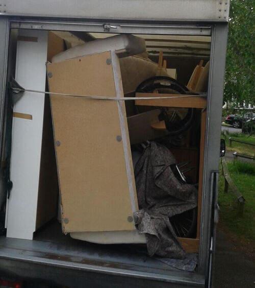 Inside a partially loaded removals van parked outdoors near a residential street, with visible houses and greenery in the background, there are various household items including a beige upholstered armchair with visible wooden legs, a dark grey fabric throw draped over part of the furniture, a light-colored wooden table or desk partially obscured behind the armchair, and a black steering wheel or similar circular object placed on top of or near the furniture. The items are stacked closely, with a wooden curb and trees visible behind the van, indicating a typical street scene in Streatham Hill, SW2. The interior of the van shows metal sides and a slide door, with a strap securing the load, suggesting a professional removals service by Man and Van Star. The scene is illuminated by natural daylight, highlighting the textures of wood, fabric, and metal, emphasizing the practical nature of house removals involving furniture and household belongings.