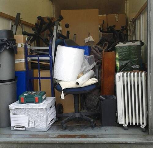 Inside a cluttered residential garage, a variety of household items and packing materials are visible. In the centre, a black office chair with a fabric seat and backrest is piled with white foam padding, several rolled sheets of bubble wrap, and a dark grey blanket. To the right of the chair, a tall wooden cabinet or wardrobe features a dark wood finish. Adjacent to it, a white portable electric oil-filled radiator heater stands upright, with its vertical fins and control panel visible. A black carry case or small luggage is positioned on the floor near the radiator. To the left of the chair, a grey cardboard moving box with a partially visible logo or label on its side sits on the concrete floor, topped with a small, open red and green toolbox. A black plastic trash bin with a lid is in the background near the left side, alongside a blue plastic container, and partially assembled exercise equipment with black frames and handlebars is visible further back. The garage's interior walls are unpainted, with exposed wooden beams and cardboard boxes stacked at the back, indicating a typical house removal preparation setting in the Streatham Hill area close to SW2 postcode, where Man and Van Star provides local removals services.
